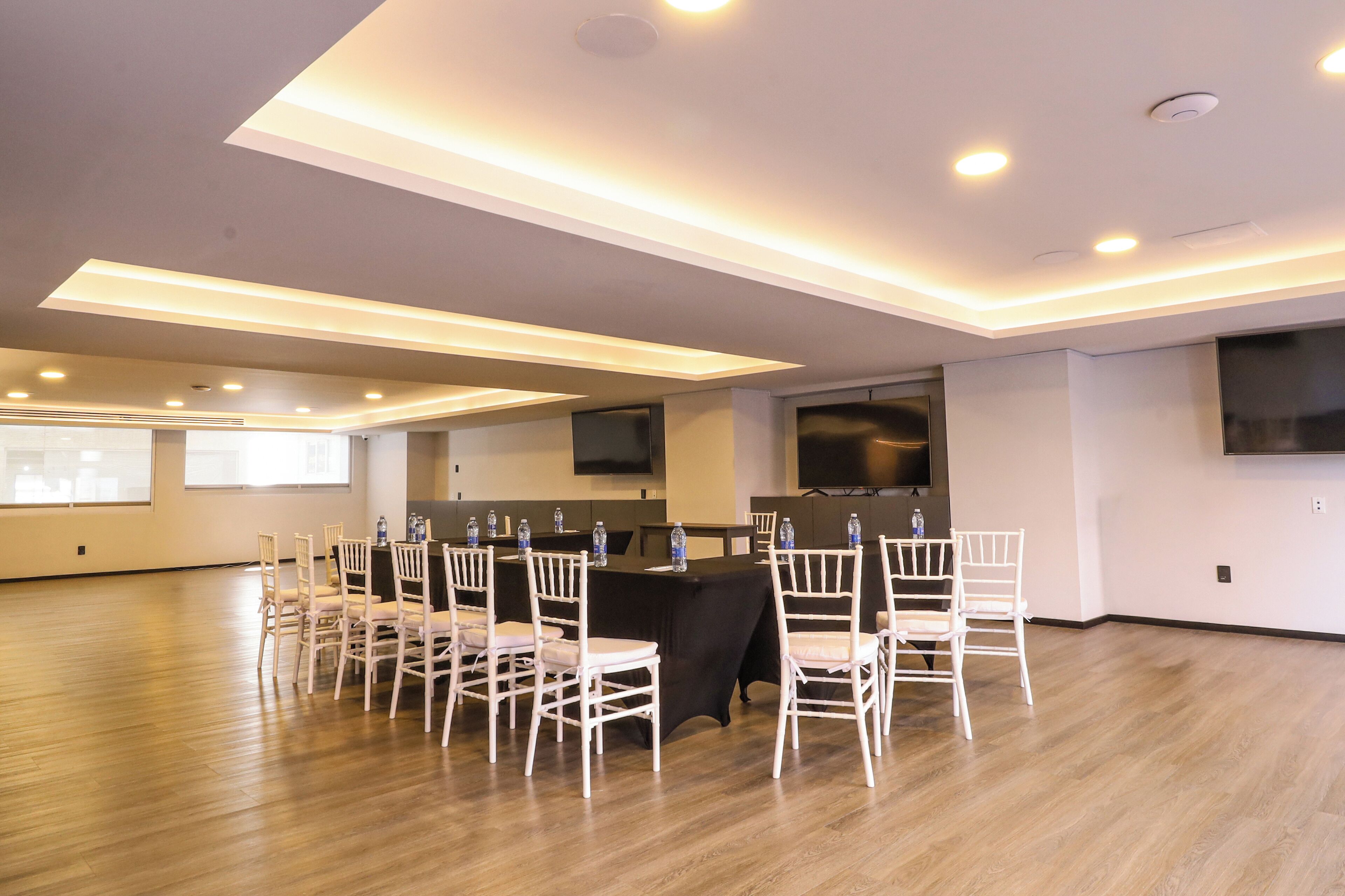 A conference room with a long table, white chairs, and a black tablecloth. There are ceiling lights and a flat-screen TV on the wall.