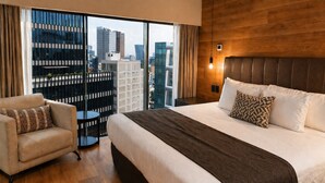 A hotel room with a large bed, a chair, a view of the cityscape through floor-to-ceiling windows, and a wooden accent wall. - St Isidro Suites Corporate Housing SPA & Wellness Center (Mexico City)