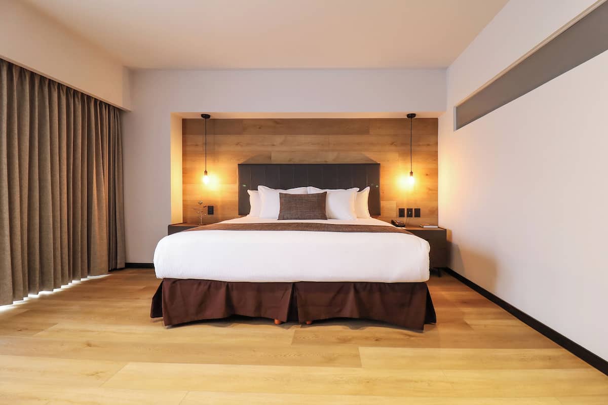 A modern hotel room with a large bed, wooden headboard, two bedside lamps, and a view of the city through the window.