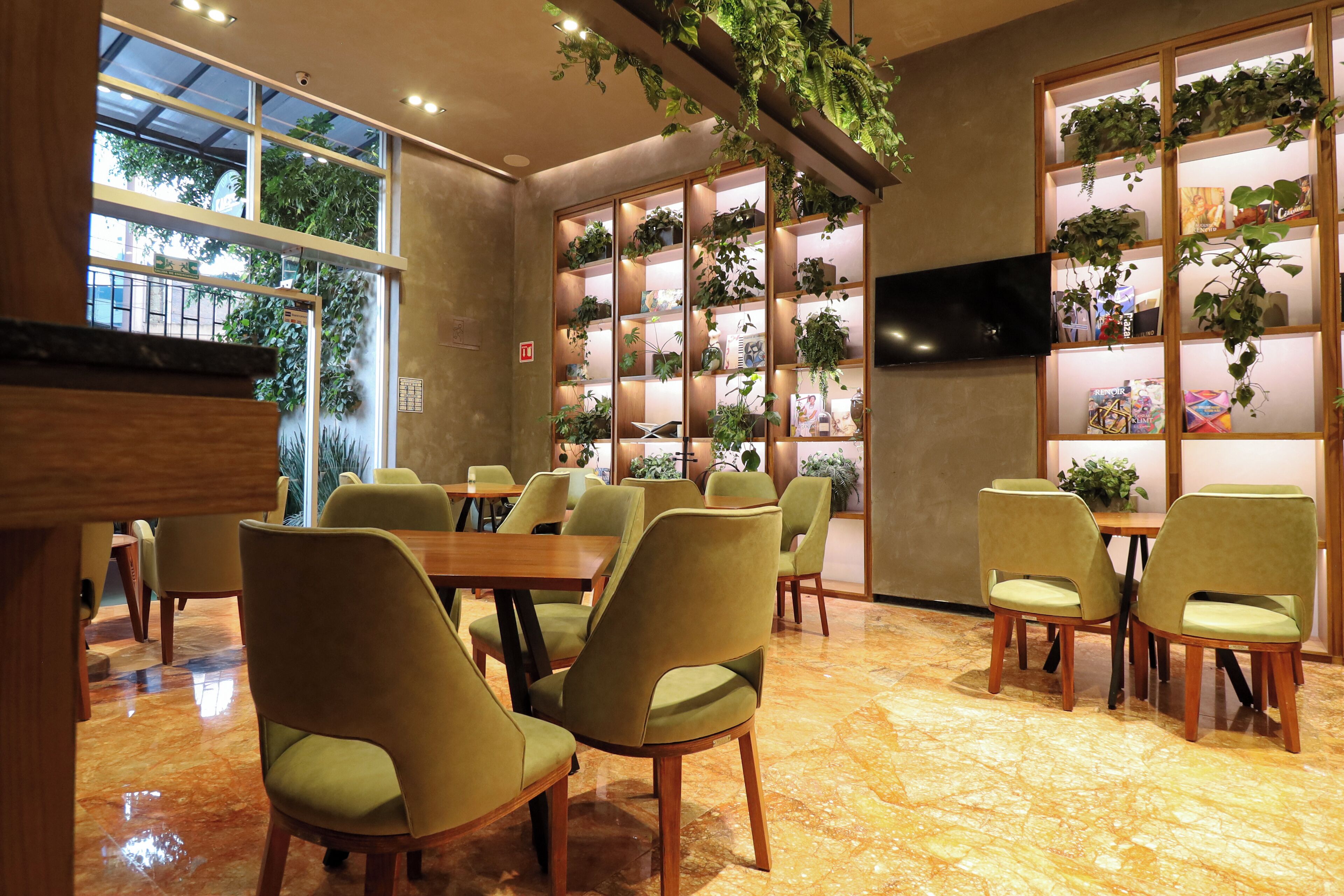 Un espace repas avec des chaises à revêtement vert, des tables en bois et un grand présentoir à plantes doté d’étagères.