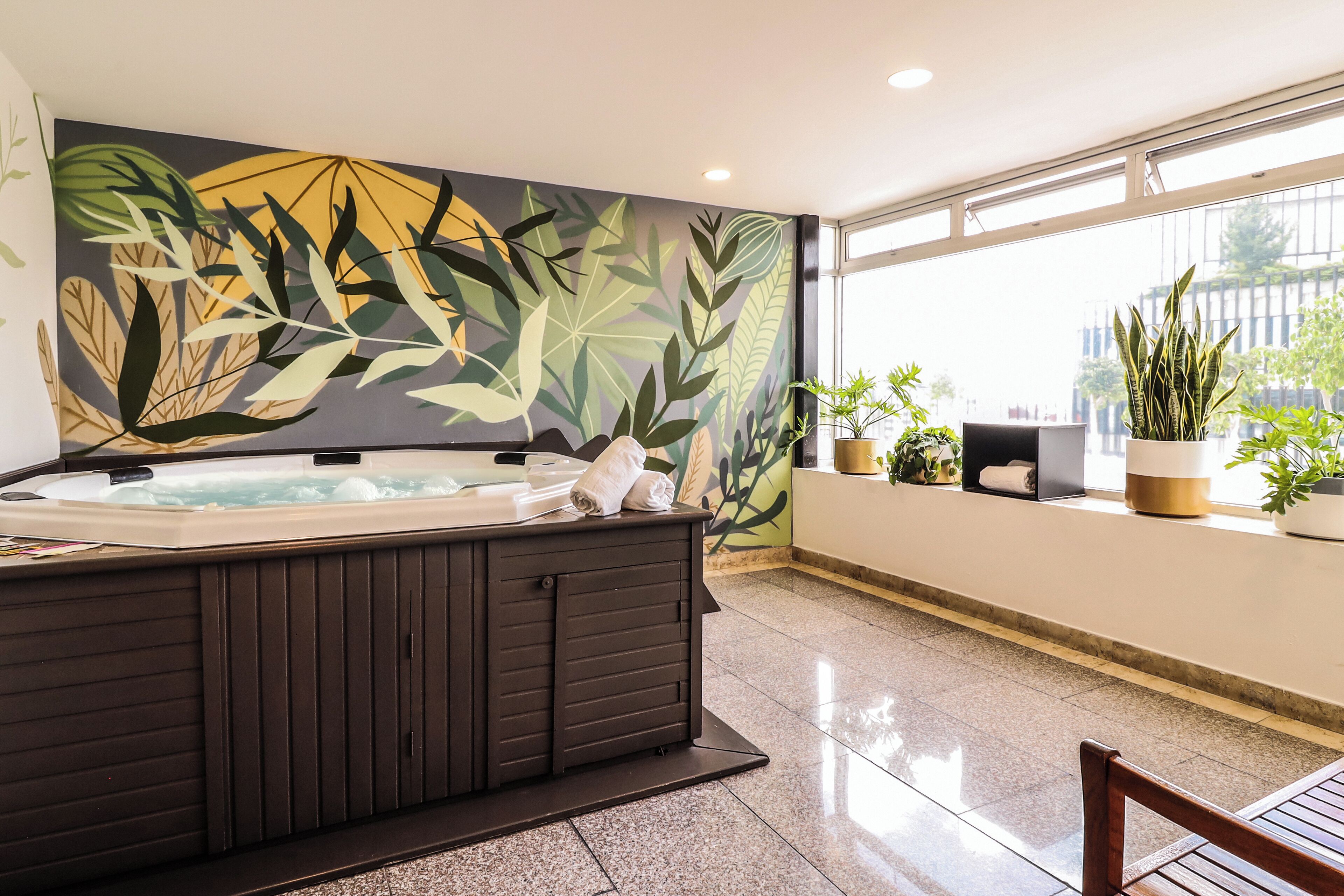 Un área de spa con una bañera de hidromasaje, un mural de hojas tropicales y plantas en macetas.