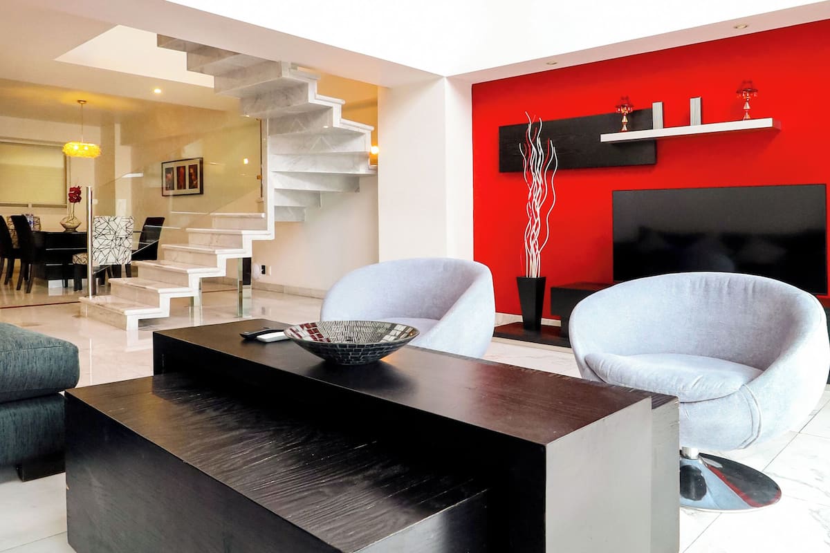 A modern living room with a black coffee table, white sofas, a red accent wall, and a staircase leading to an upper level.