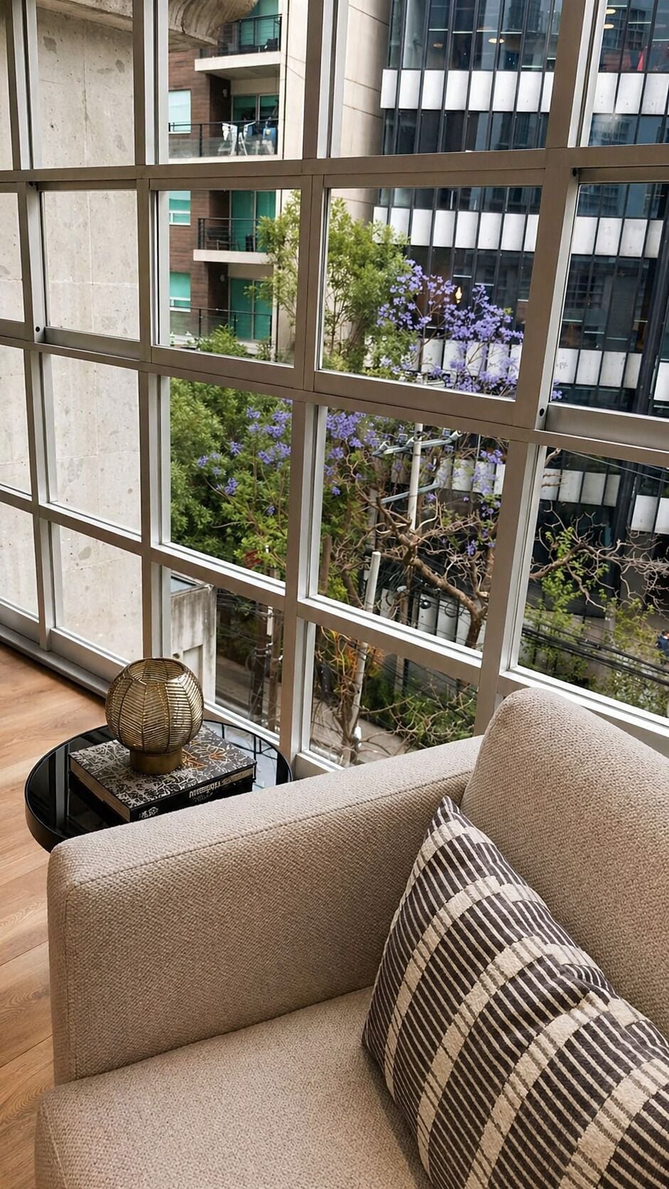 Um interior moderno com um sofá, um abajur decorativo e vista para um edifício com varandas e árvores.