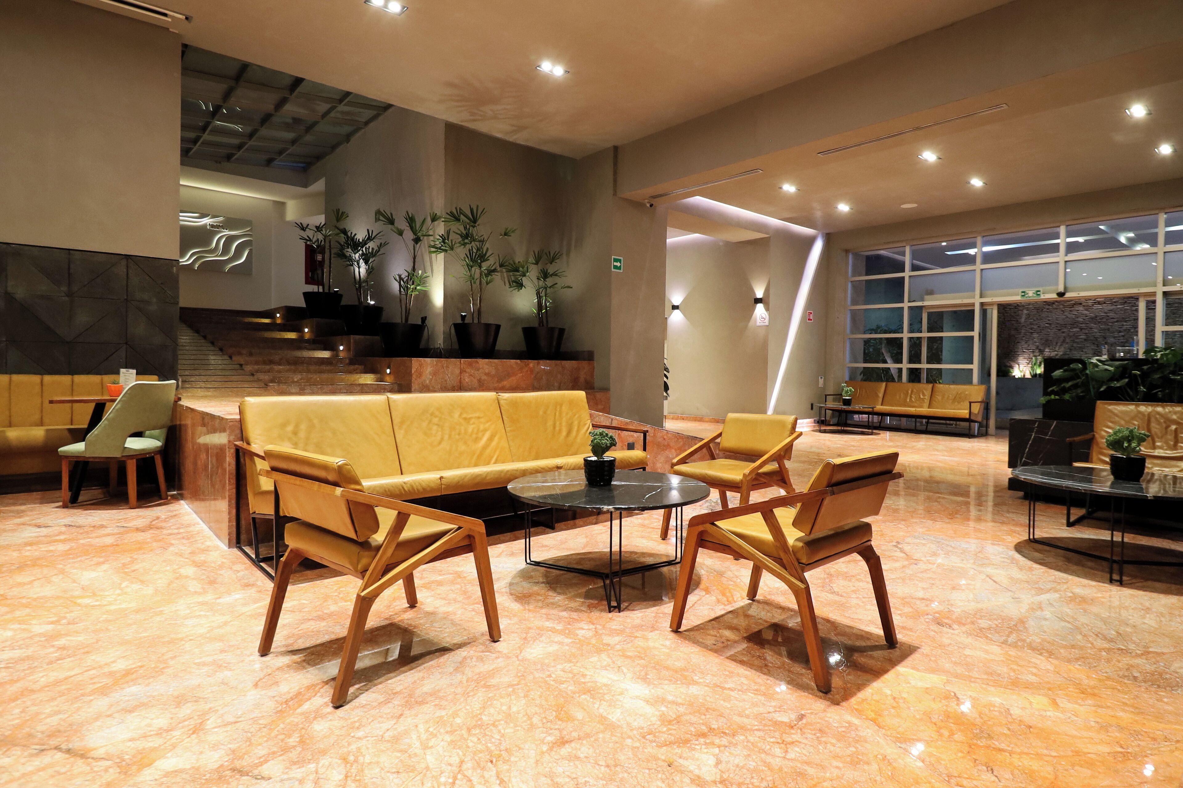 A modern lobby with a yellow sofa, wooden chairs, a marble floor, and potted plants.