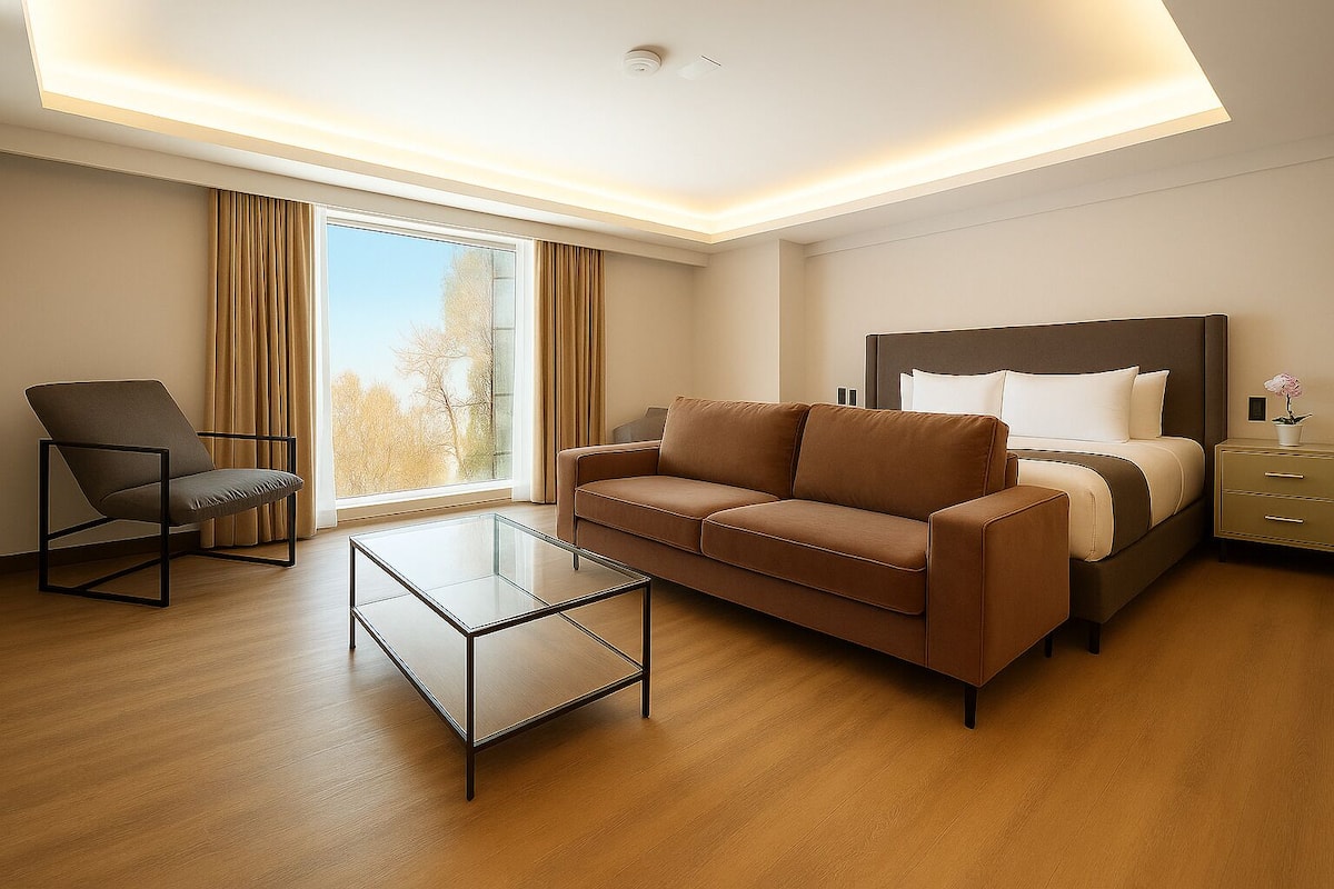 A modern hotel room with a brown sofa, a glass coffee table, a chair, a bed with a headboard, and a nightstand.