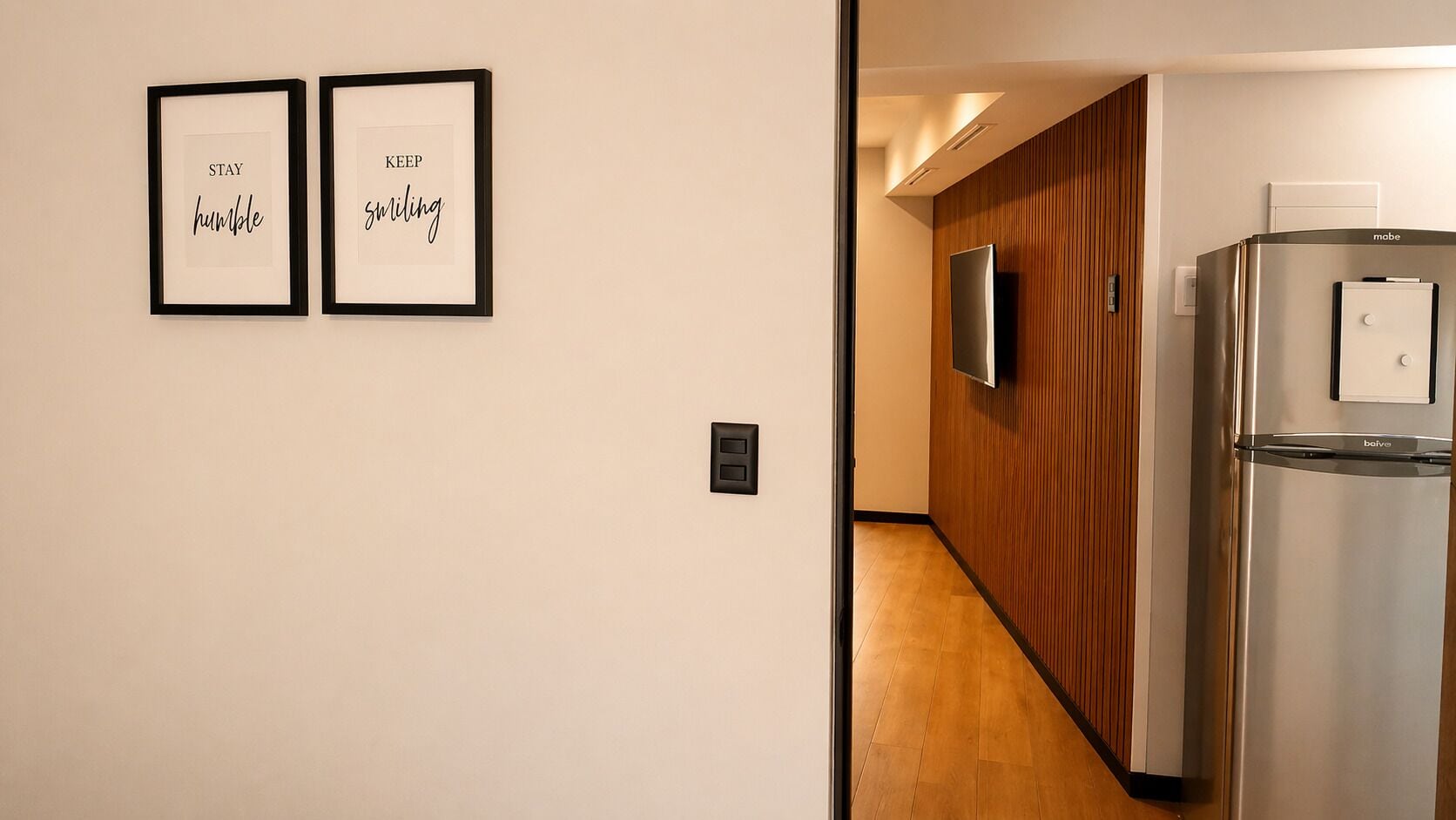 A hallway with two framed motivational quotes, a wooden wall, a stainless steel refrigerator, and a door leading to another room.