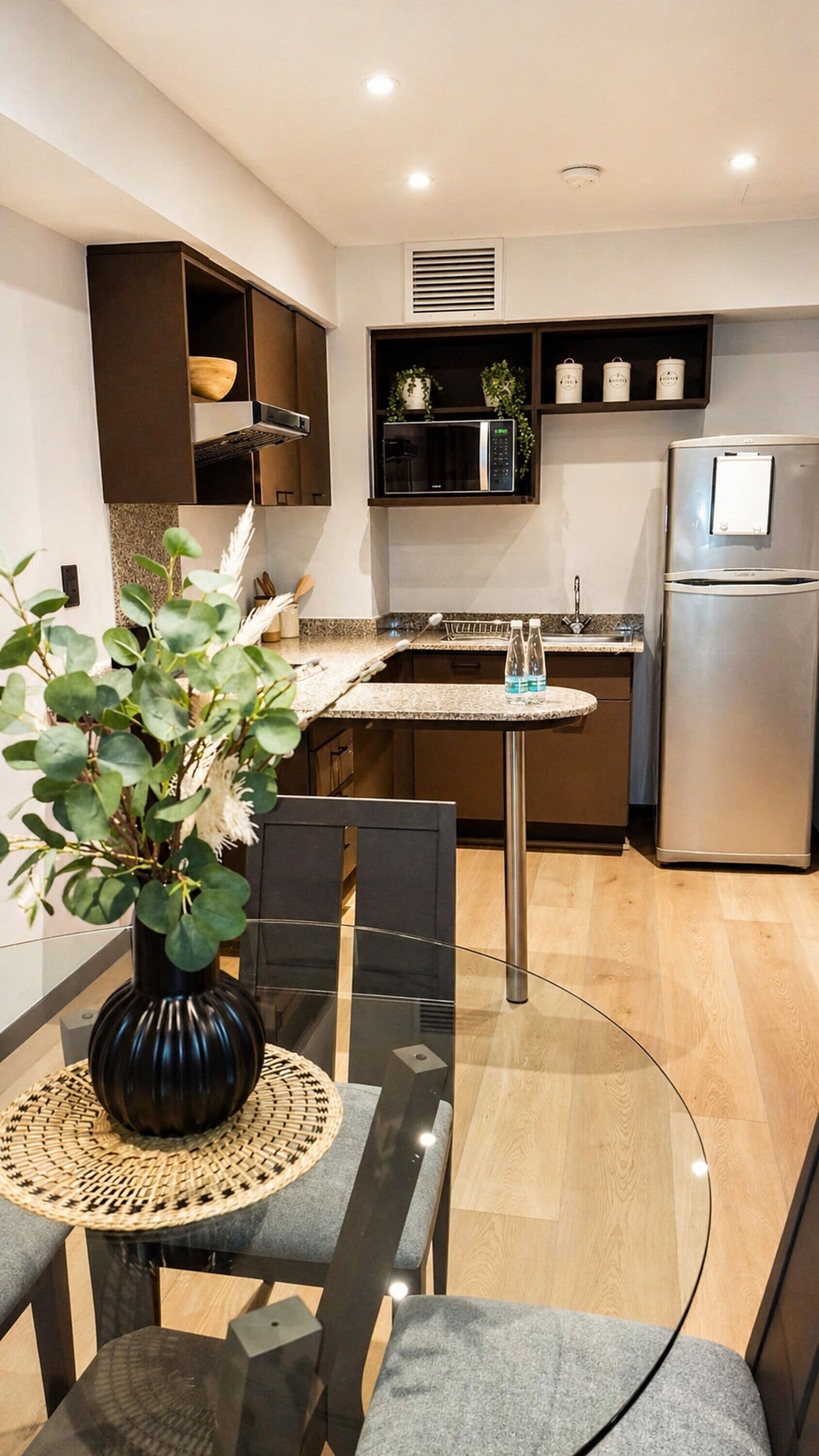 Une cuisine moderne avec une table à manger en verre, des chaises, un vase noir avec des feuilles vertes, un four à micro-ondes et un réfrigérateur.