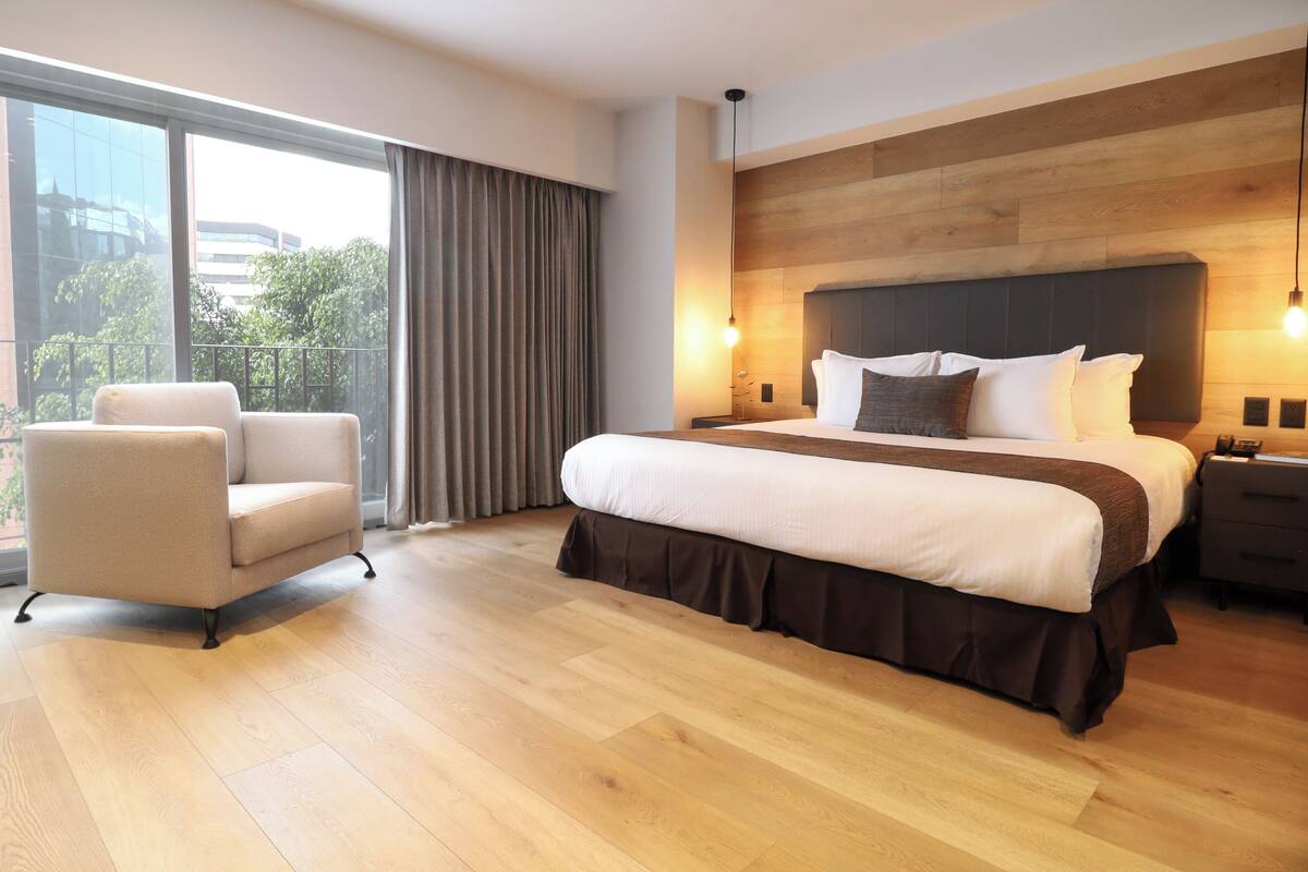 A modern hotel room with a large bed, a chair, and a view of the city through large windows.