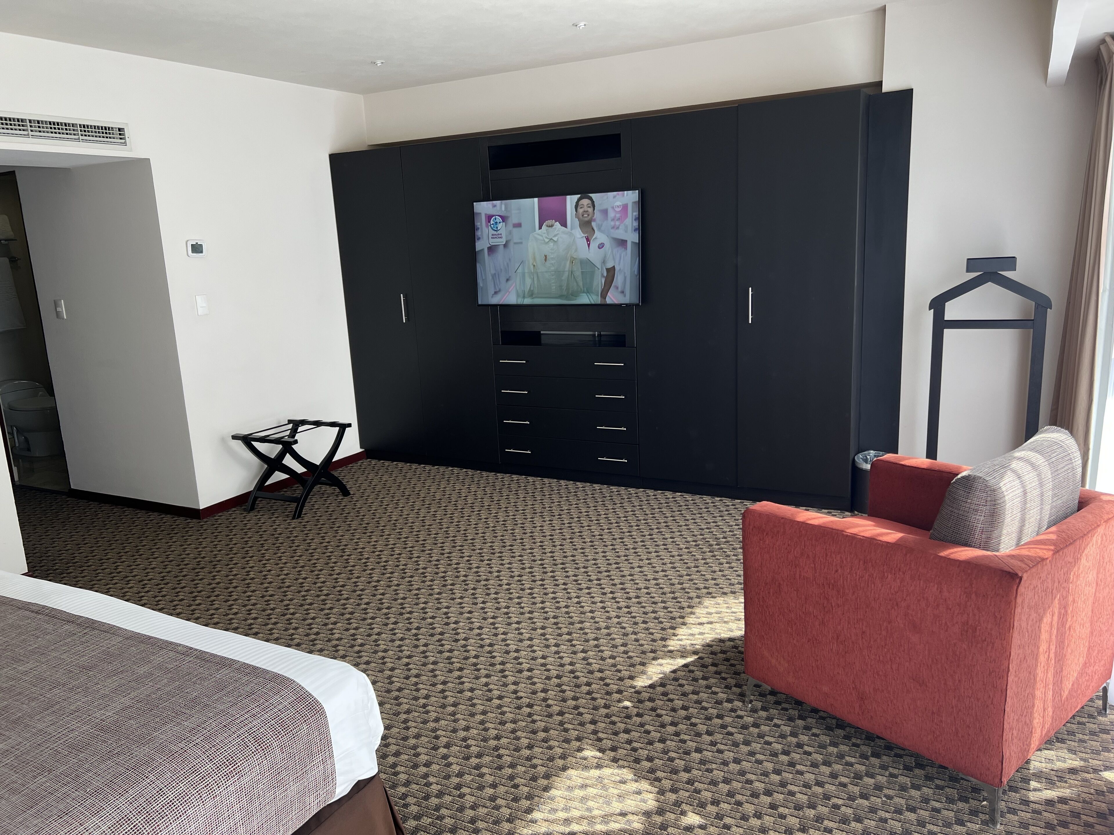 A hotel room with a large wardrobe, a television mounted on the wall, a red armchair, and a patterned carpet.
