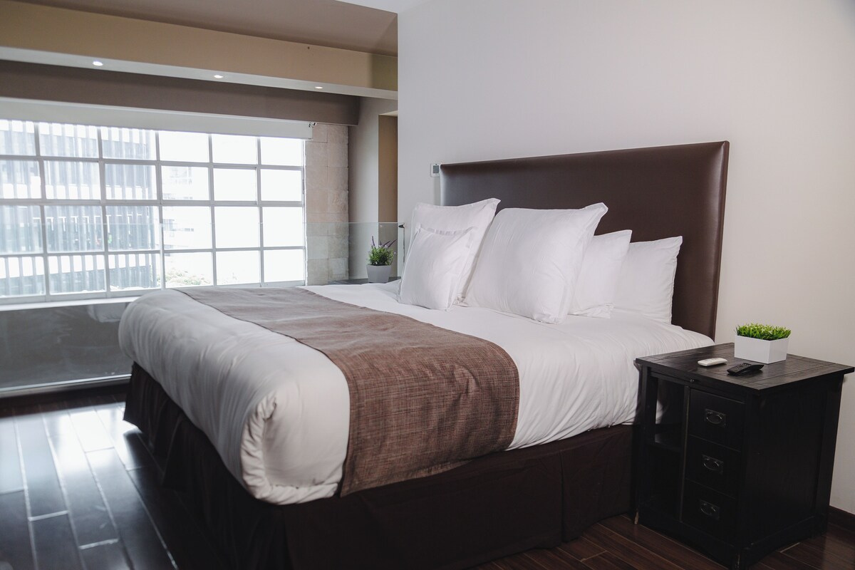 A modern hotel room with a large bed, a nightstand, a potted plant, and a window with a city view.