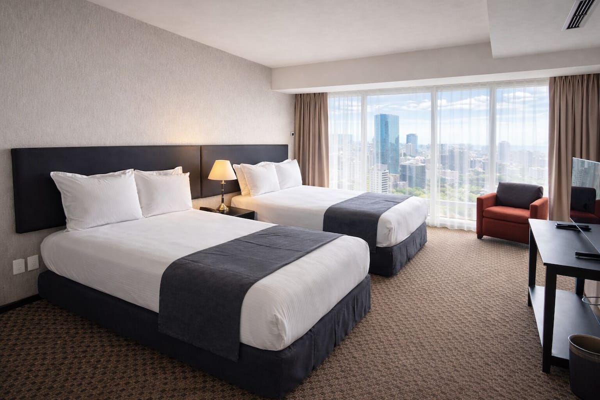 A hotel room with two beds, a desk, a chair, and a large window with a city view.