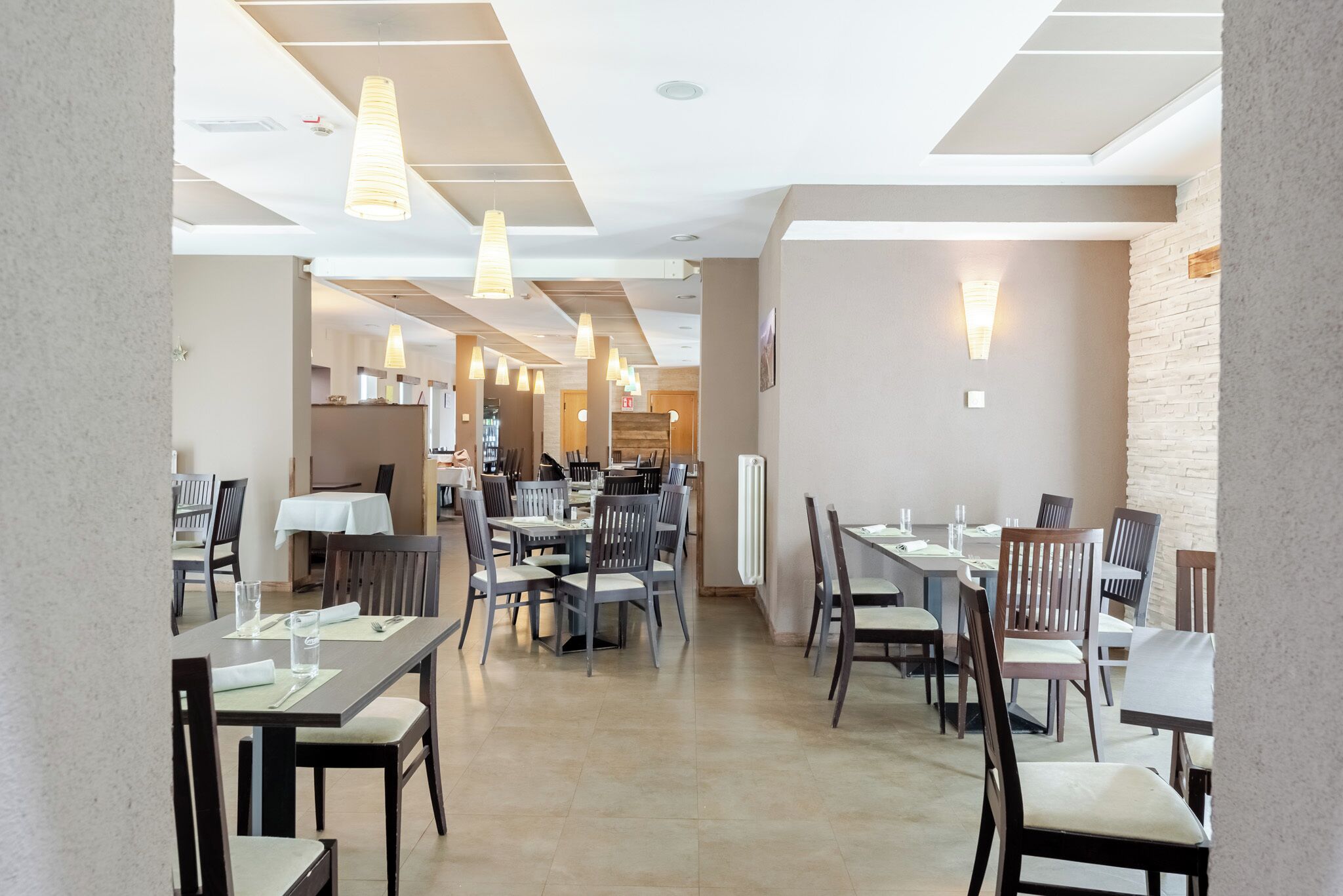 A modern dining area with multiple tables and chairs, overhead lighting, and a kitchen in the background.