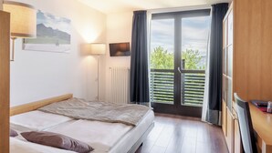 Quarto de hotel com cama, escrivaninha, cadeira, televisão, luminária e varanda com vista para o verde.