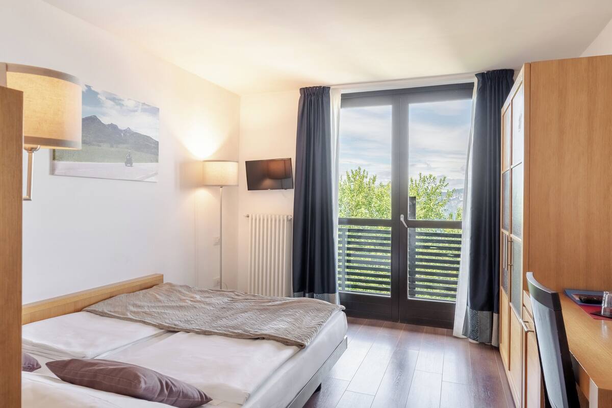 Een hotelkamer met een bed, bureau, stoel, televisie, lamp en balkon met uitzicht op groen.