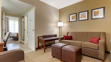 A hotel room with a sofa, ottoman, and a small table. There is a lamp, framed pictures on the wall, and a door leading to another room.