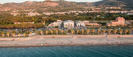 Vista aérea de un pueblo costero con playa, edificios y montañas al fondo.