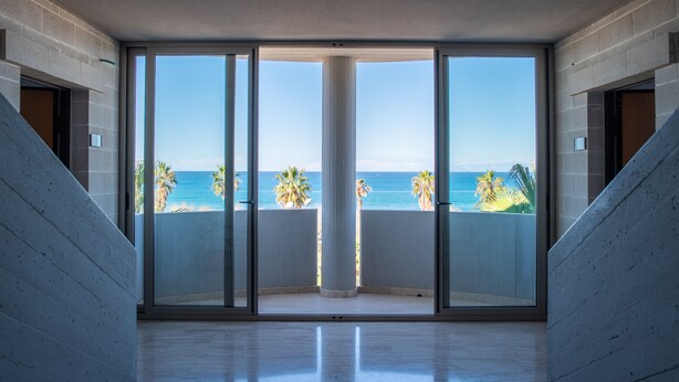 Een ruime kamer met een grote glazen deur die uitkomt op een balkon met uitzicht op de oceaan en palmbomen.