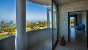 Een balkon met uitzicht op de oceaan en palmbomen, een potplant, en een blauwe bank in de aangrenzende kamer.