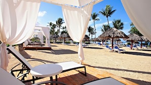 A beachside lounge area with white canopies, lounge chairs, and thatched umbrellas.