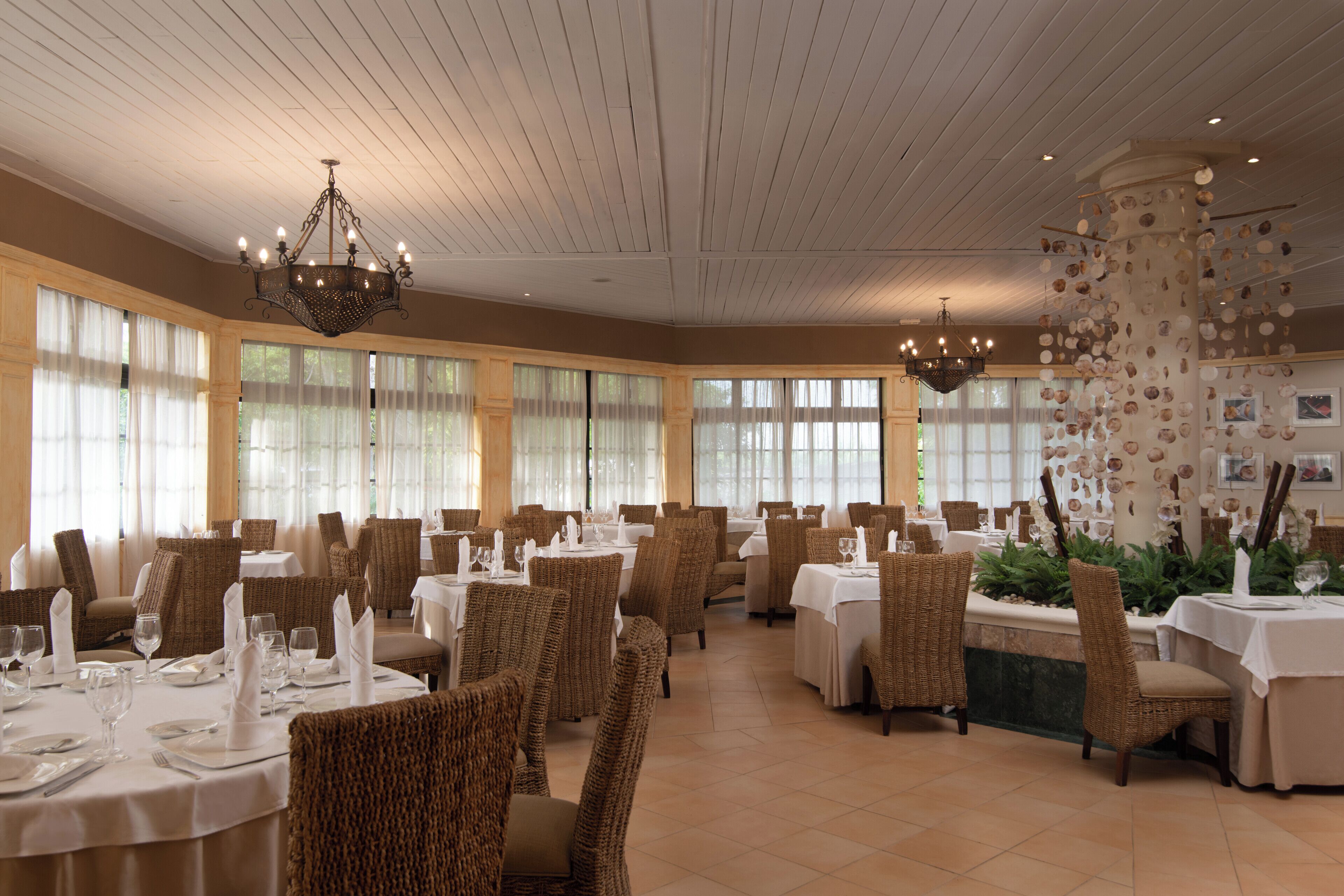 Un restaurant avec des tables rondes, des chaises en osier, des lustres et de grandes fenêtres.