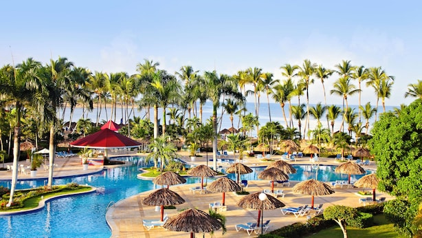 A resort pool area with palm trees, lounge chairs, and a red-roofed pavilion.