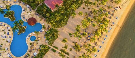 Luchtbeeld van een resort met meerdere gebouwen, een zwembad en een strandgebied met ligbedden.