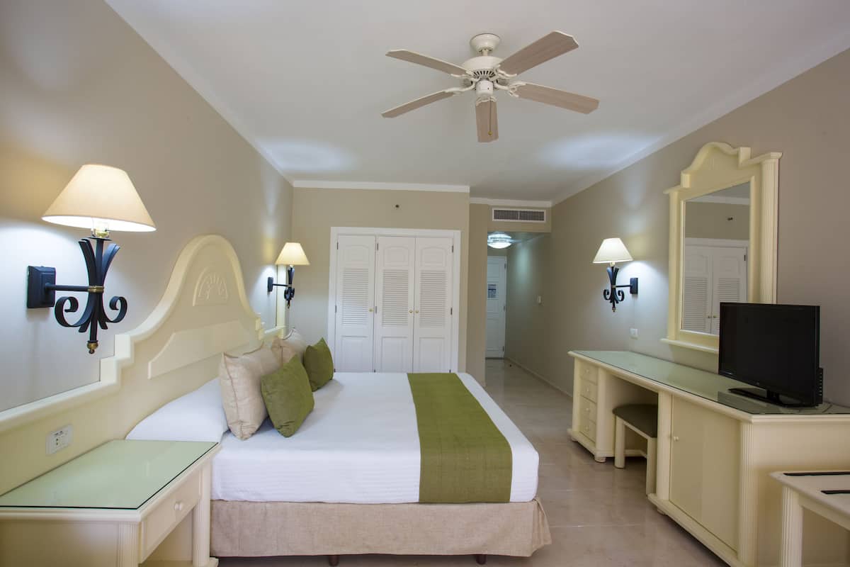 Une chambre d’hôtel avec un lit, des lampes de chevet, un bureau avec un ordinateur, un ventilateur de plafond et un grand miroir.