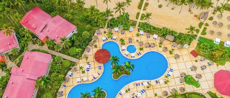 Eine Vogelperspektive auf ein Resort mit einem zentralen Schwimmbad, Strandliegen und Palmen.