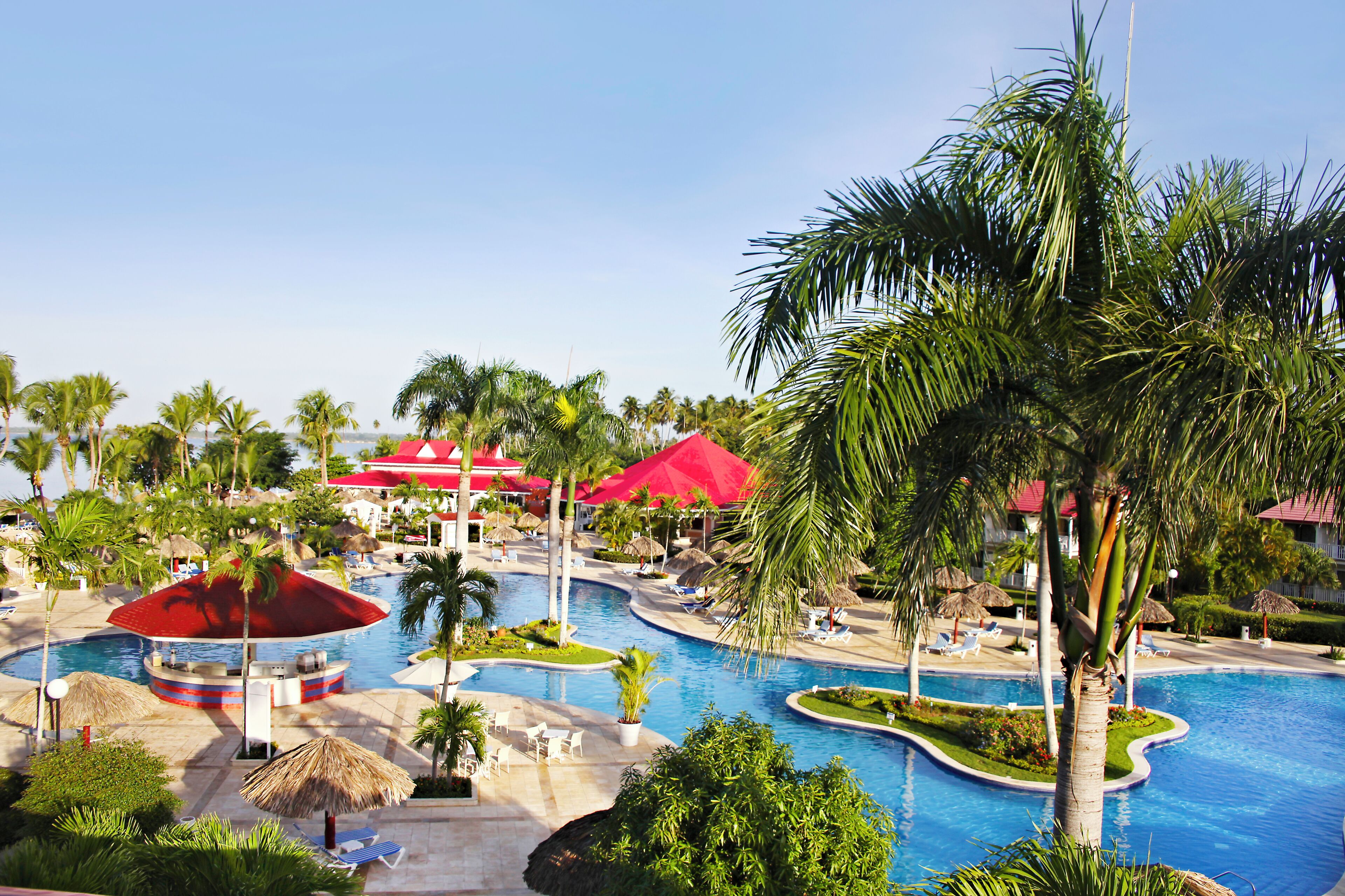 Un resort con piscina central, sillas de descanso, palmeras y edificios con techo rojo.