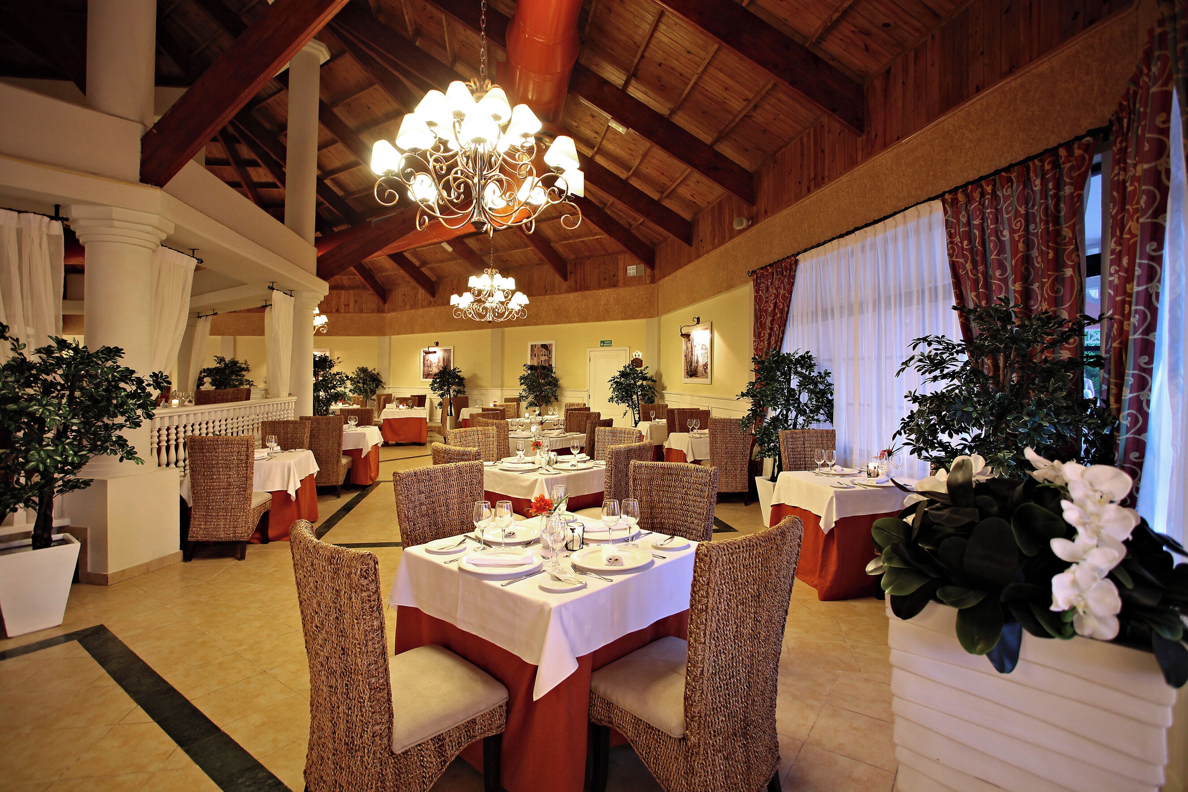 Un restaurant avec de hauts plafonds, des lustres et de nombreuses tables, toutes recouvertes de nappes blanches et entourées de chaises.