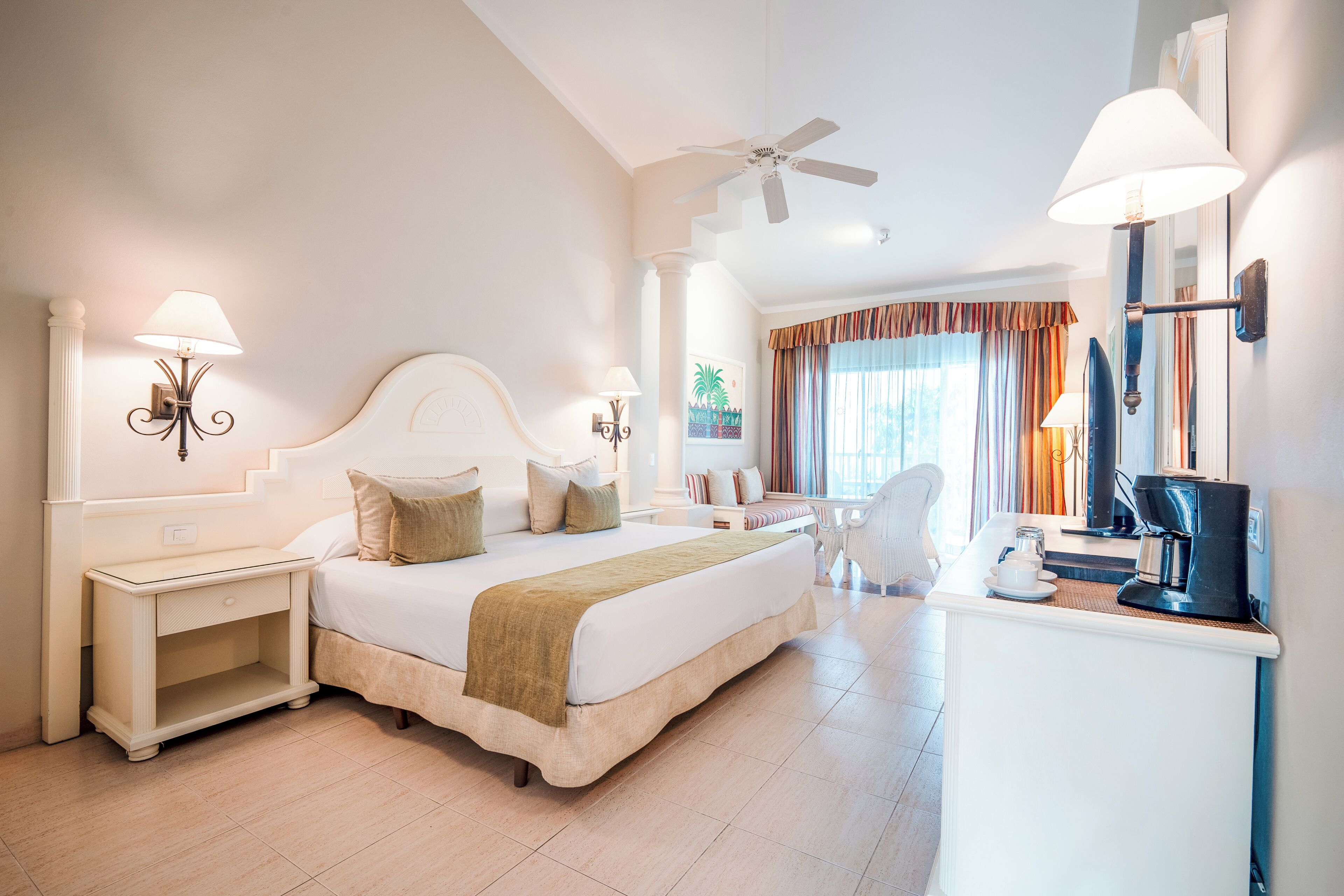 Una camera d'albergo con un letto grande, una scrivania, una sedia, una ventola a soffitto e una macchina per il caffè.