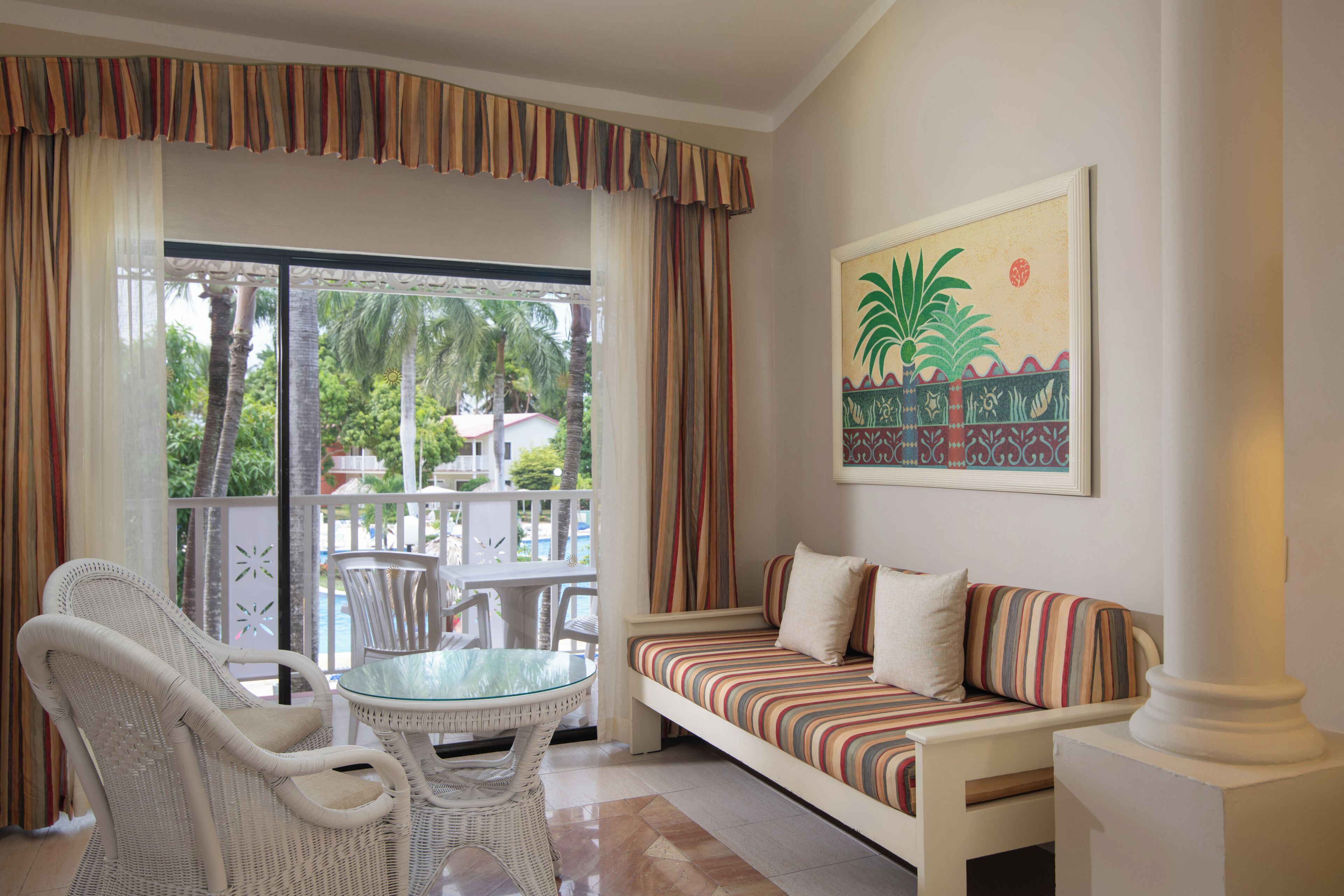 A room with a sofa, chairs, a glass table, and a view of a pool and palm trees.