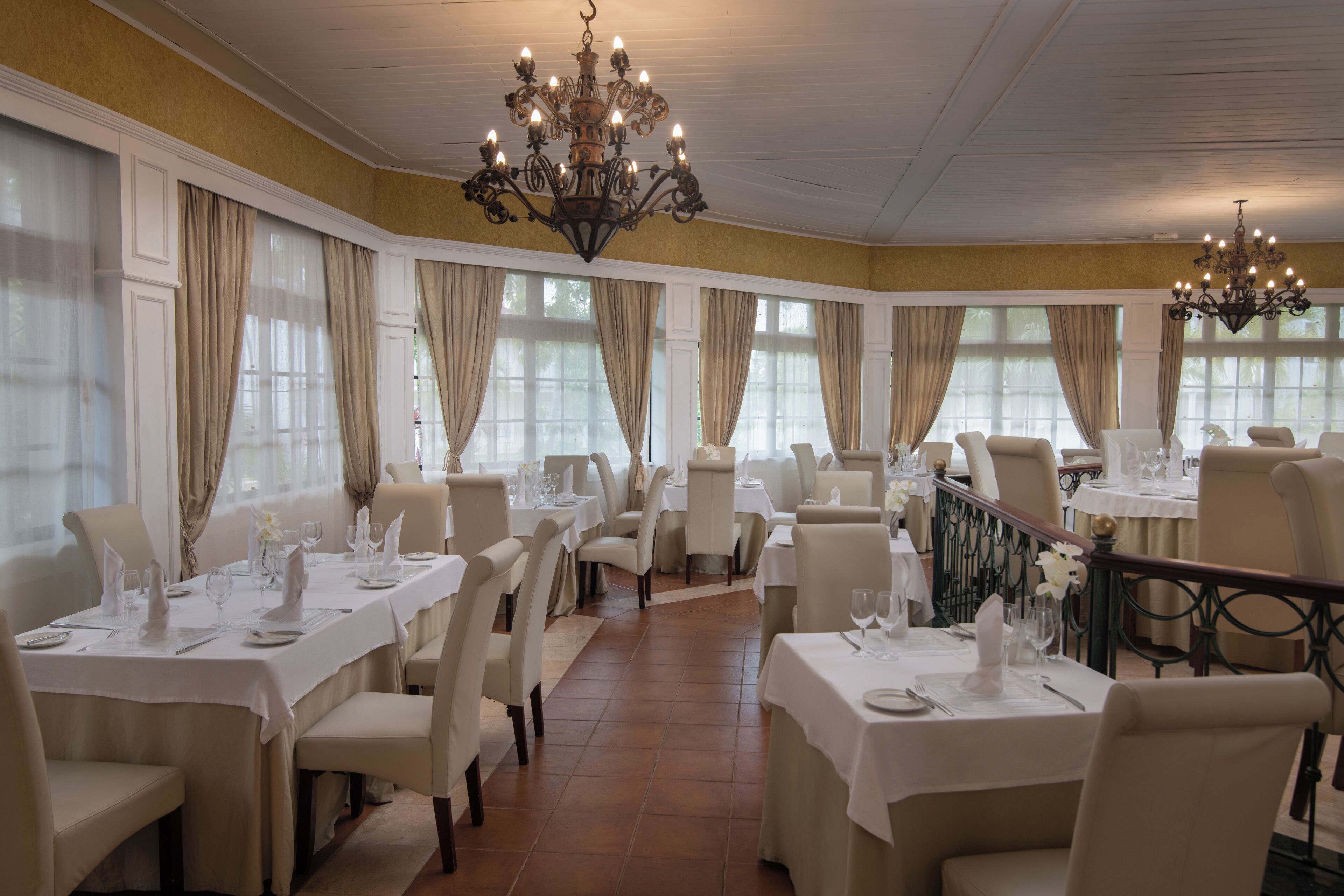 A restaurant with multiple tables set for dining, chandeliers, and large windows with curtains.