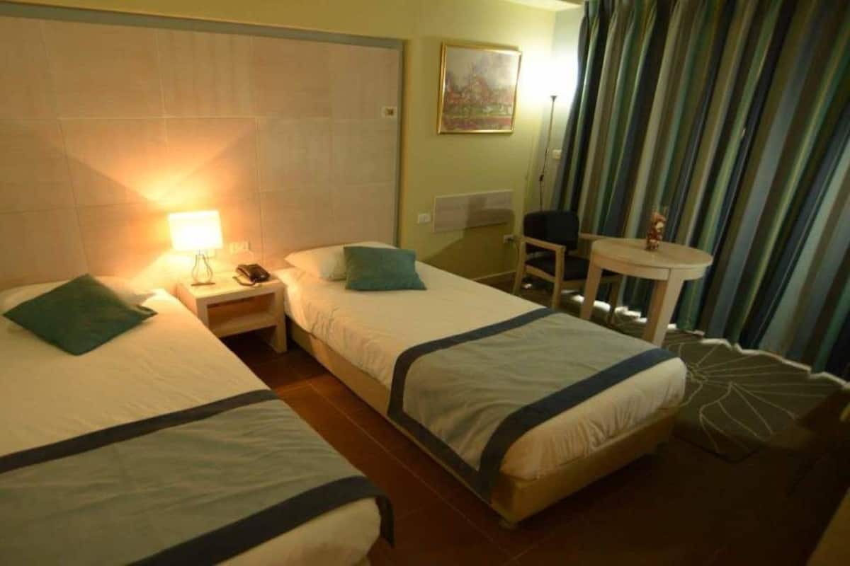 A hotel room with two single beds, a small table, a chair, a lamp, and a window with curtains.
