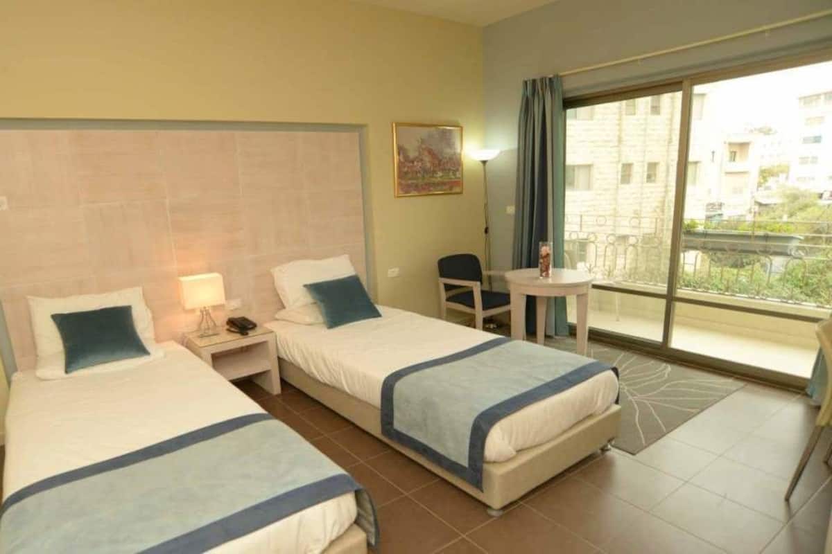 A hotel room with two beds, a small table, a chair, and a balcony with a view.