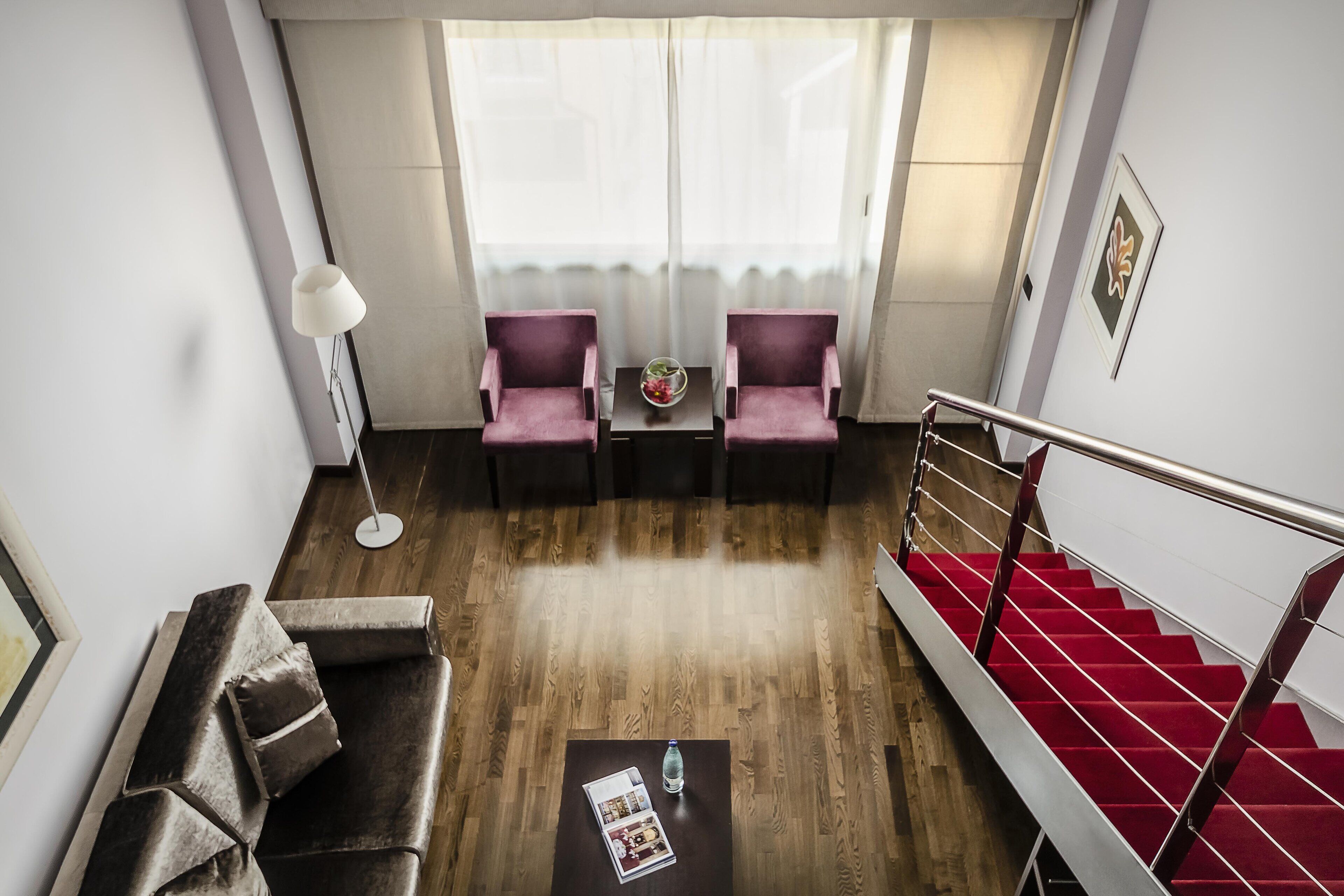 A modern hotel room with a wooden floor, a red carpeted staircase, a sofa, two armchairs, a small table with a vase, and a large window with sheer curtains.