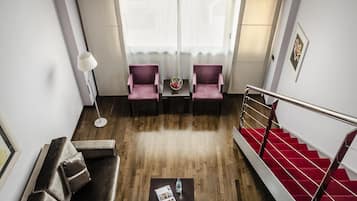 A modern hotel room with a wooden floor, a red carpeted staircase, a sofa, two armchairs, a small table with a vase, and a large window with sheer curtains.