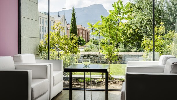 Un espace extérieur moderne, aménagé avec des canapés blancs et une table en verre, offrant une vue sur un jardin et des bâtiments situés au loin.