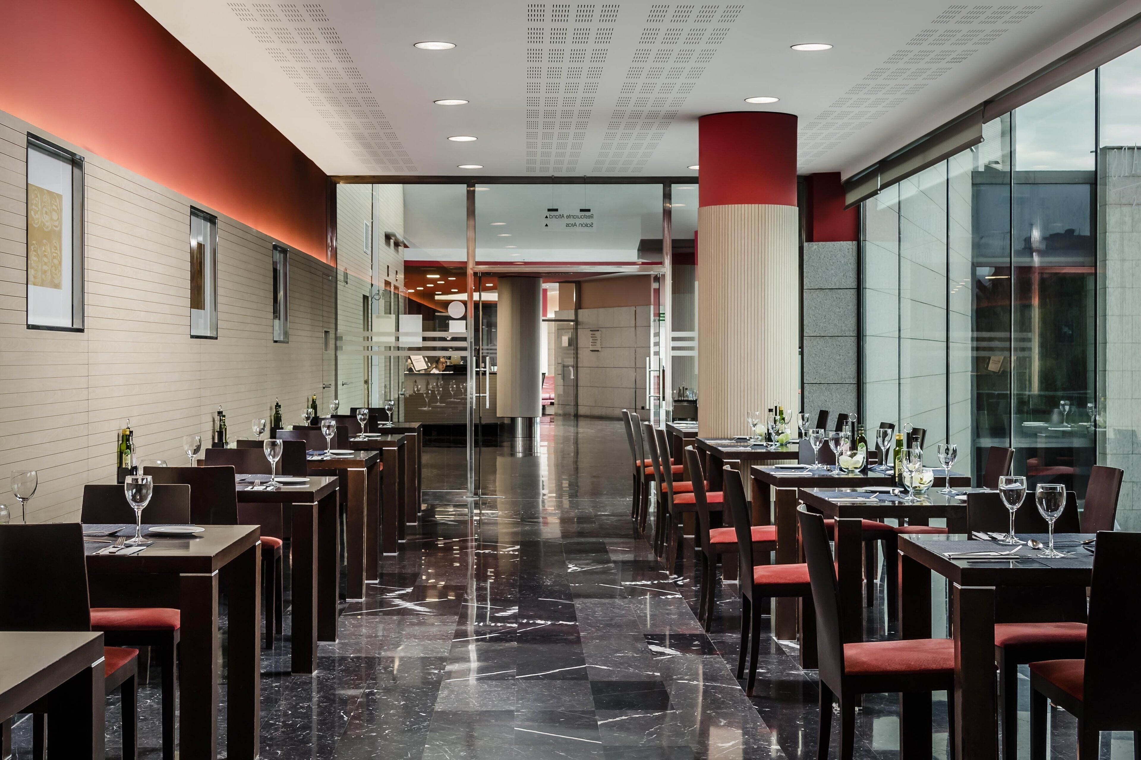 Un restaurante moderno con suelo de mármol pulido, paredes blancas y rojas, y separadores de vidrio. Las mesas están dispuestas con copas de vino y cubiertos.
