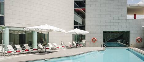 Ein Schwimmbecken mit Liegestühlen und Sonnenschirmen, umgeben von einem Gebäude mit einer Kachelfassade.