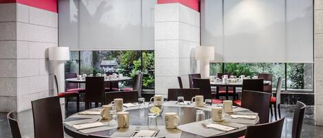 Un comedor moderno con mesas redondas, sillas con acolchado rojo y ventanas amplias que dan a vistas de vegetación.