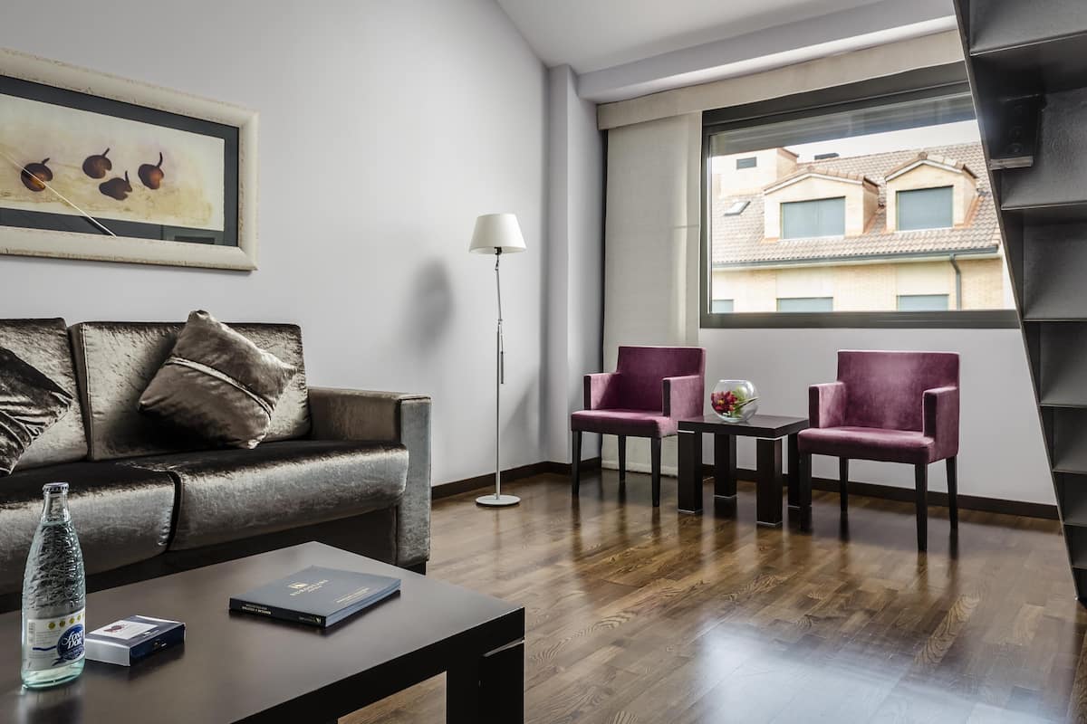 Een moderne woonkamer met een donker leren bank, twee paarse fauteuils, een salontafel en een vloerlamp. Een lijstwerk met kunst hangt aan de muur, en een raam biedt uitzicht op gebouwen.
