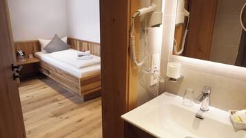 Una habitación de hotel con piso de madera, una cama con cabecera, una mesita de noche con lámpara, un espejo, un baño con lavamanos, una toalla y un secador de pelo.