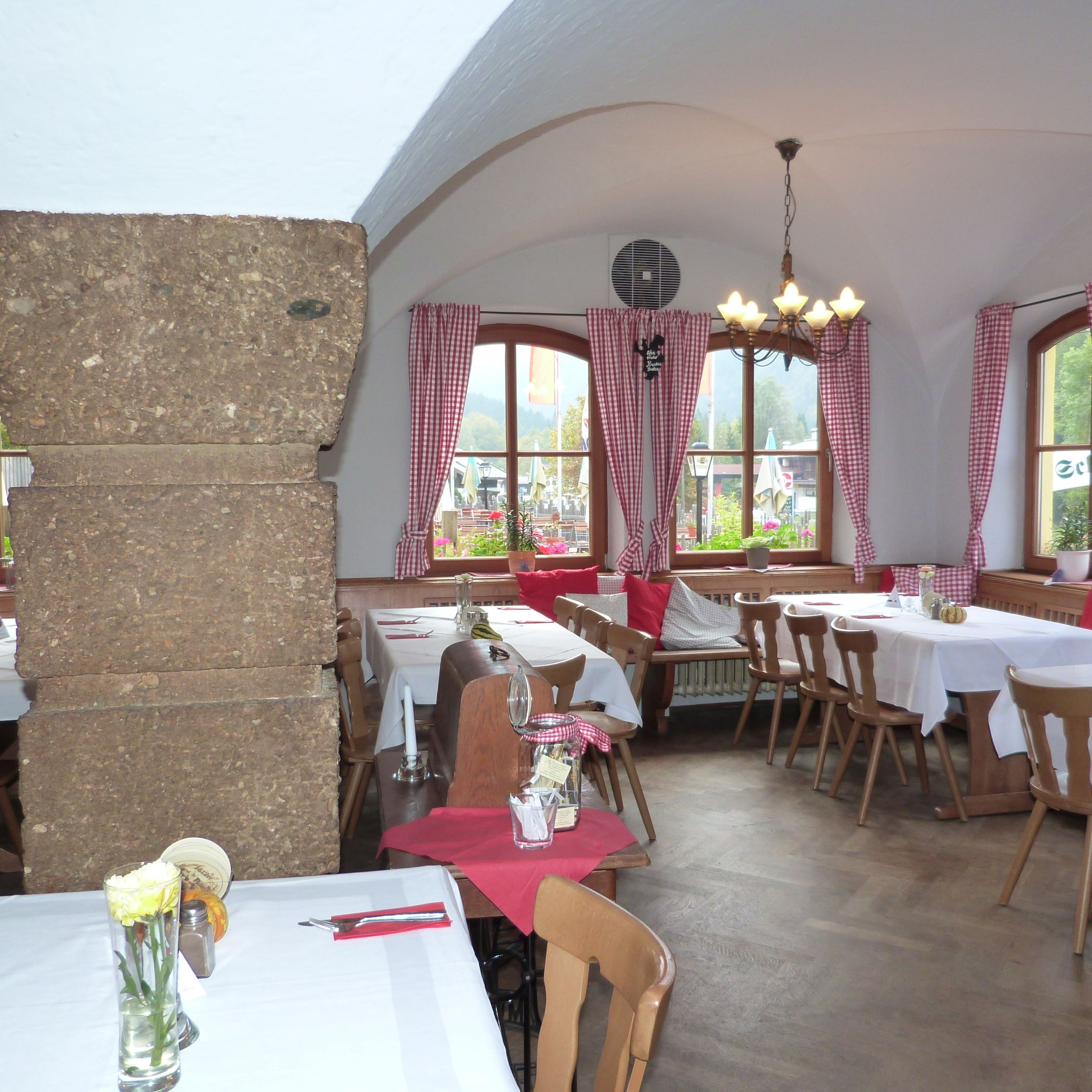 Un restaurante con varias mesas y sillas, ventanas amplias con cortinas a cuadros y una pared de piedra en un lateral.