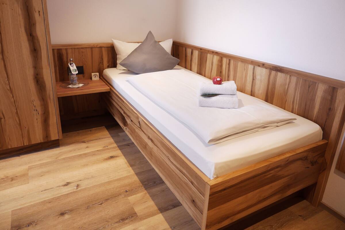 A wooden bunk bed with a white mattress and pillows, a small wooden nightstand with a lamp, and a wooden floor.