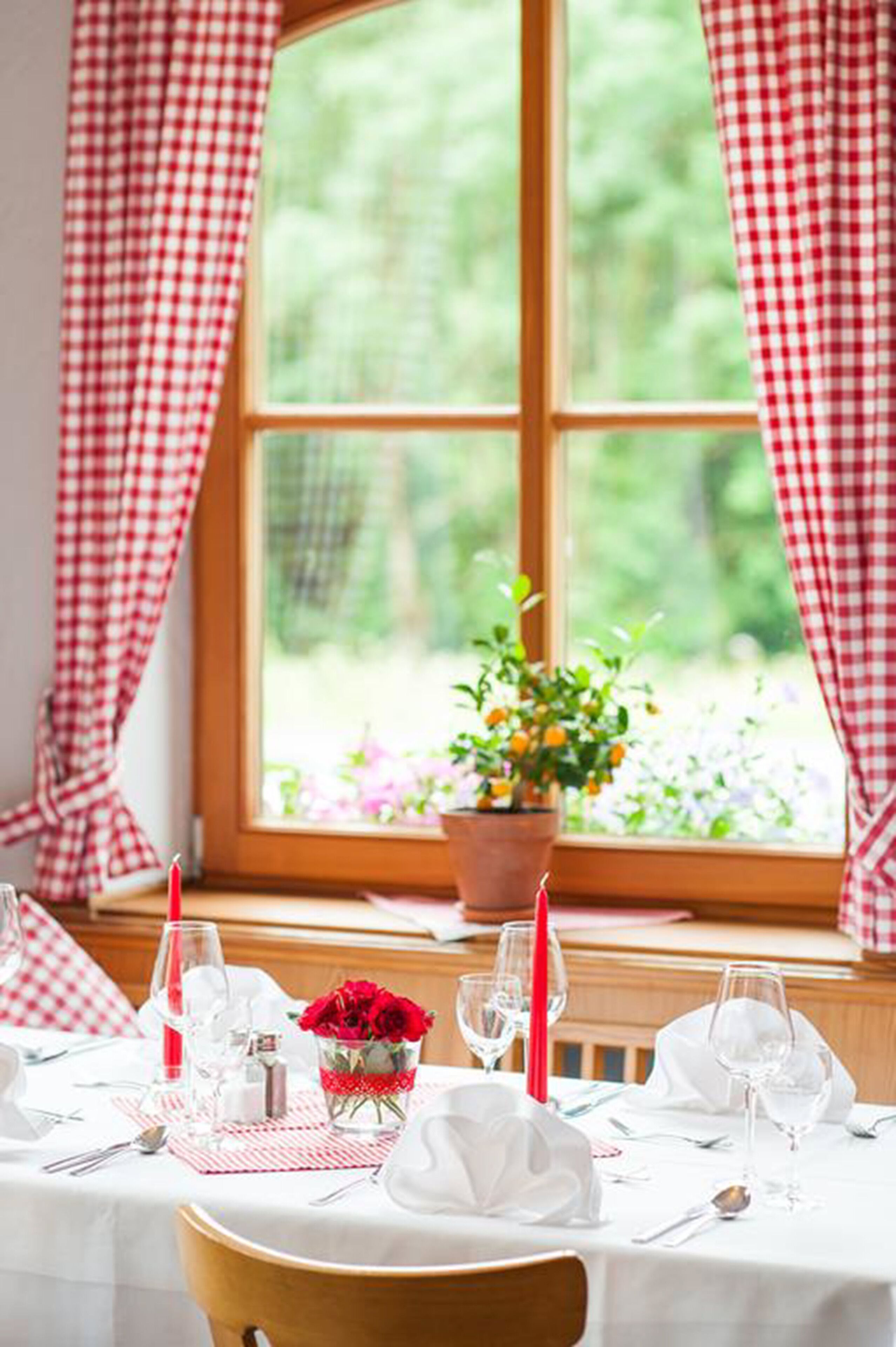 Una mesa de comedor para dos, con una planta en maceta y flores rojas en el alféizar de la ventana.