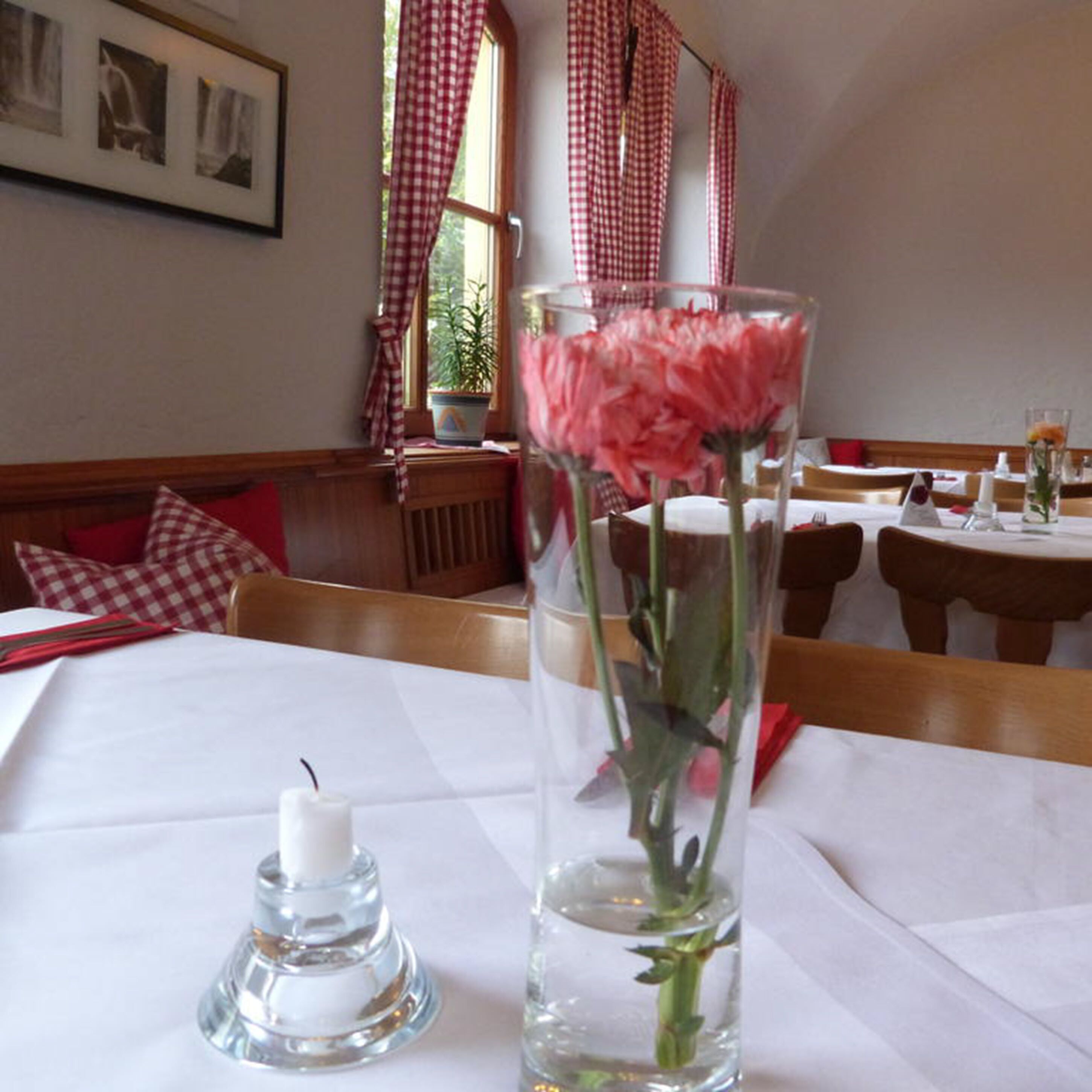Ein Esstisch mit Kerze, einer Glaskaraffe mit Blumen und karierten Vorhängen in einem Restaurant.