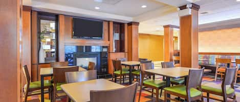A hotel dining area with tables and chairs, a television mounted on the wall, and a patterned carpet.