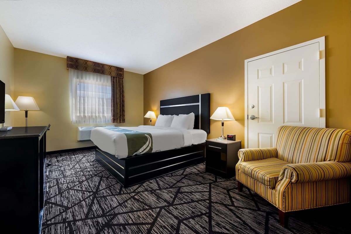 A hotel room with a bed, two bedside tables with lamps, a sofa, a window with curtains, and a door.