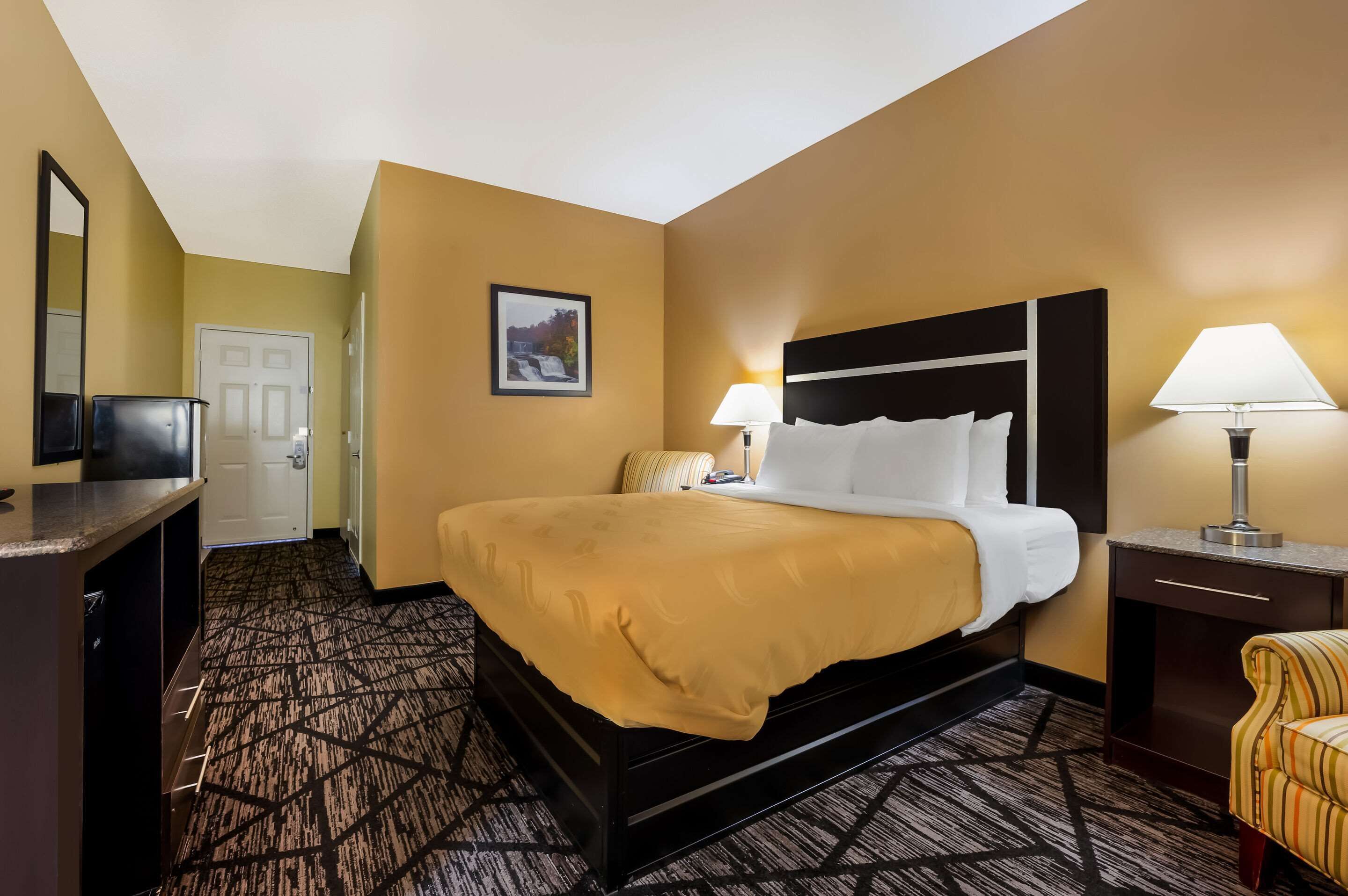 Une chambre d’hôtel avec un grand lit, un bureau, une chaise, un miroir, une porte et une lampe fixée au mur.